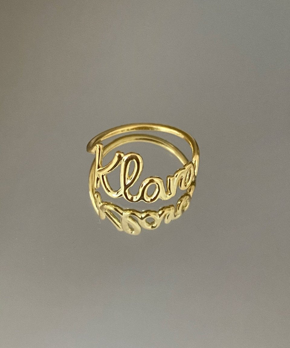 A gold-colored personalized ring with the name 'Klara' written on it, displayed against a gray background.