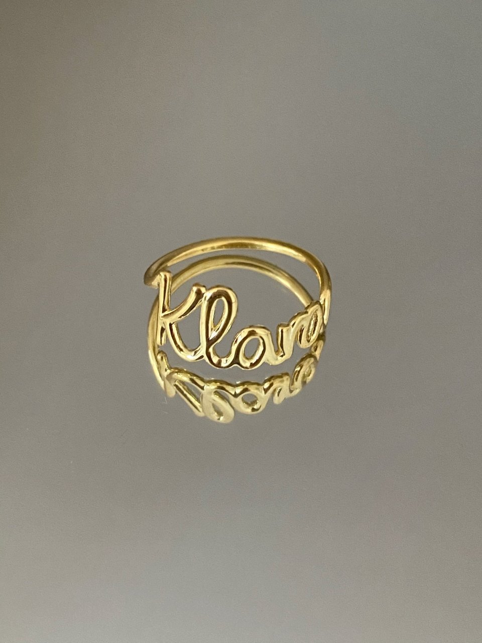 A gold-colored personalized ring with the name 'Klara' written on it, displayed against a gray background.
