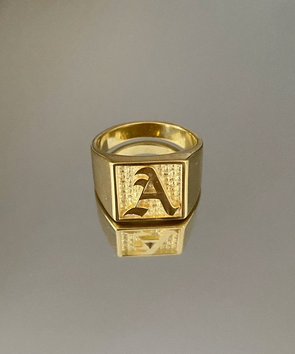 A gold-colored square signet ring with an engraved letter 'A' on it, displayed against a gray background.