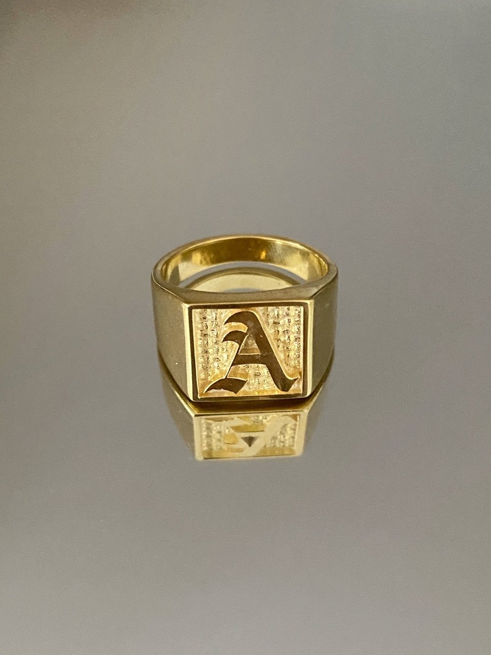 A gold-colored square signet ring with an engraved letter 'A' on it, displayed against a gray background.