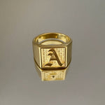 A gold-colored square signet ring with an engraved letter 'A' on it, displayed against a gray background.