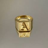 A gold-colored square signet ring with an engraved letter 'A' on it, displayed against a gray background.