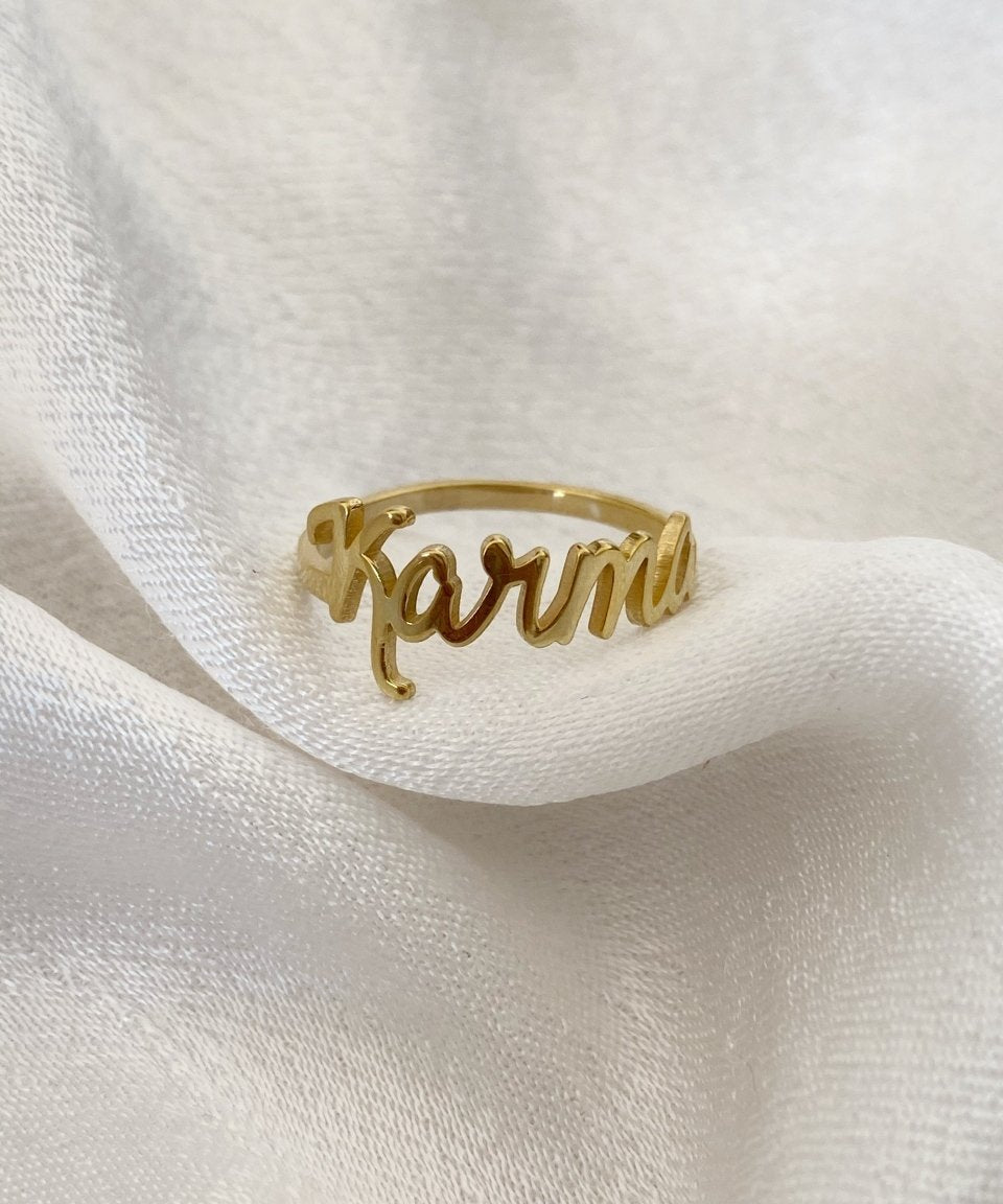 A gold-colored custom ring with the name 'Karma' written on it, displayed on a white fabric surface.
