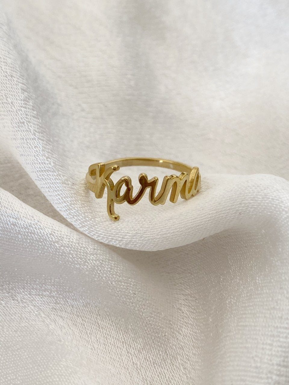 A gold-colored custom ring with the name 'Karma' written on it, displayed on a white fabric surface.